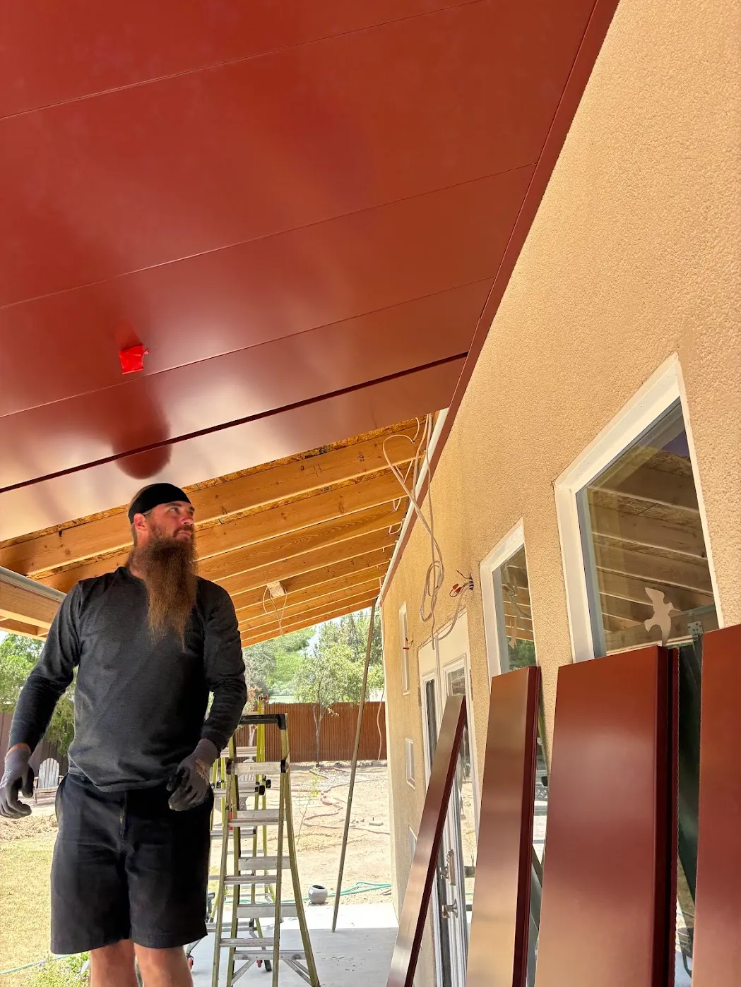 Installer under a freshly completed red standing seam patio cover, additional panels staged for the next run