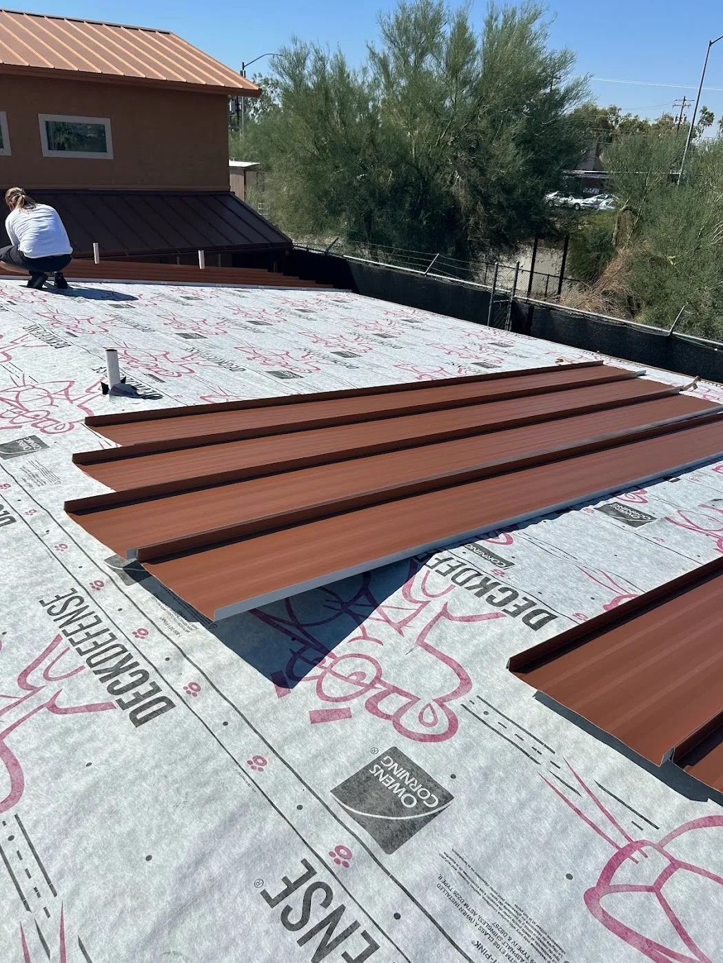 Copper-tone standing seam panels laid out on a roof deck over Owens Corning DeckDefense underlayment, installer fastening a run